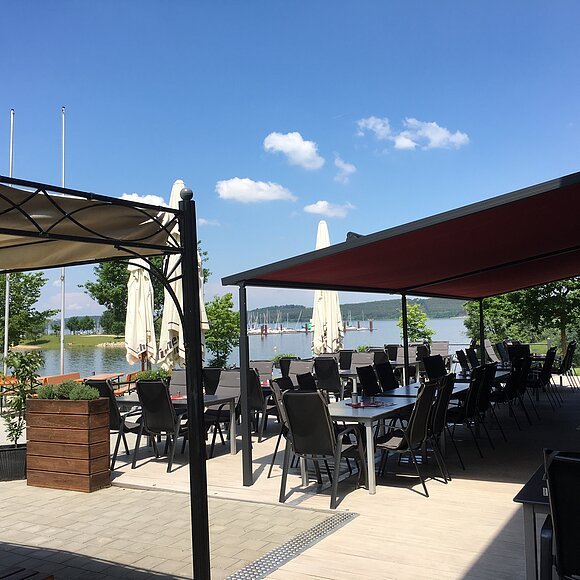 Die Bucht Eine große geplasterte Terasse am See. Im Vordergrund eine Markisse und ein Pavillon. Schwarze Stühle und Tische stehen in der Sonne.