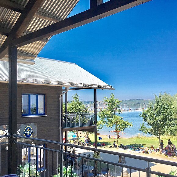 Die Bucht am Brombachsee Blick von einem Balkon auf ein zweistöckiges Holzhaus mit Terasse. Dahinter eine Liegewiese mit See.