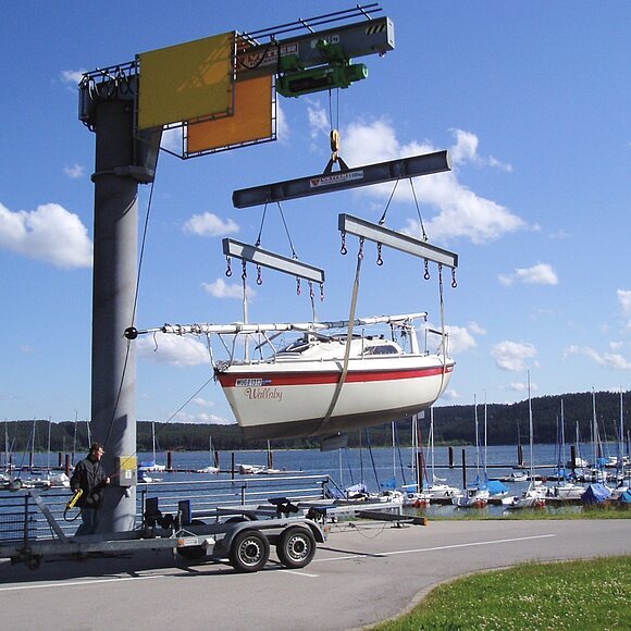 Ein Segelboot wird mit der Hilfe Bootskran auf den Anhänger eines Autos geladen. Das rot-weiße Boot hängt in der Luft. Ein Mann bedient den Kran.
