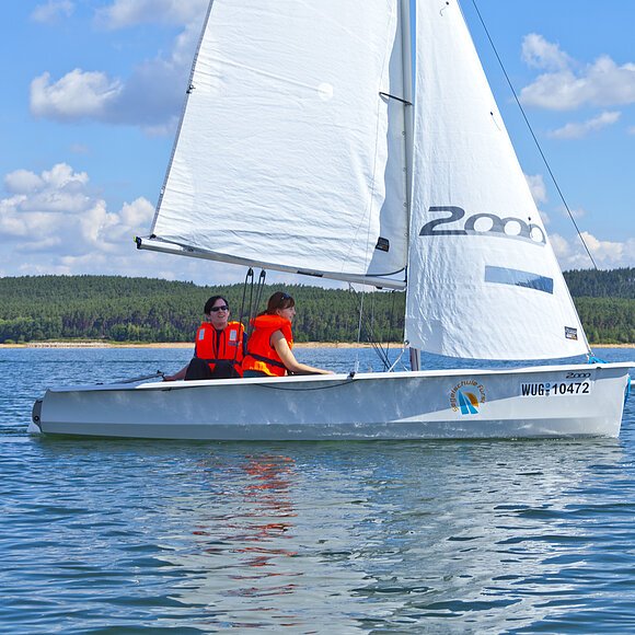 Segelschule Fürst Zwei Segler mit oranger Warnweste fahren mit ihrem weißen Segelboot über den See. Am Ufer im Hintergund erstreckt sich ein grüner Wald unter blauem Himmel.