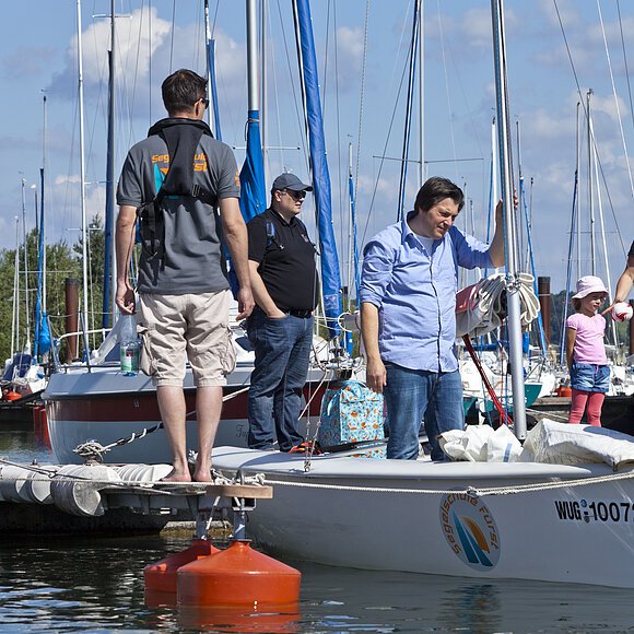 Auf Probe stellen Urlauber stehen auf Steg eines Segelboothafens. Eine Frau vernkoten die Seile eines Segelboot. Ein Mann schaut ihr dabei zu. Links von ihm steht ein Mann mit Schwimmweste mit dem Rücken zur Kamera. Dahinter stehen noch vier weiter Urlauber am Steg.