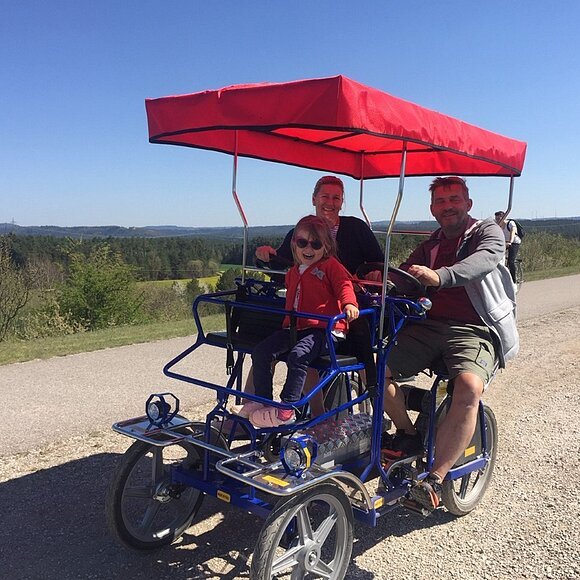 Eine Ehepaar mit Kind fährt auf einer Rikscha auf einem Schotterweg durch eine grüne Landschaft. Die Familie lächelt in die Kamera.