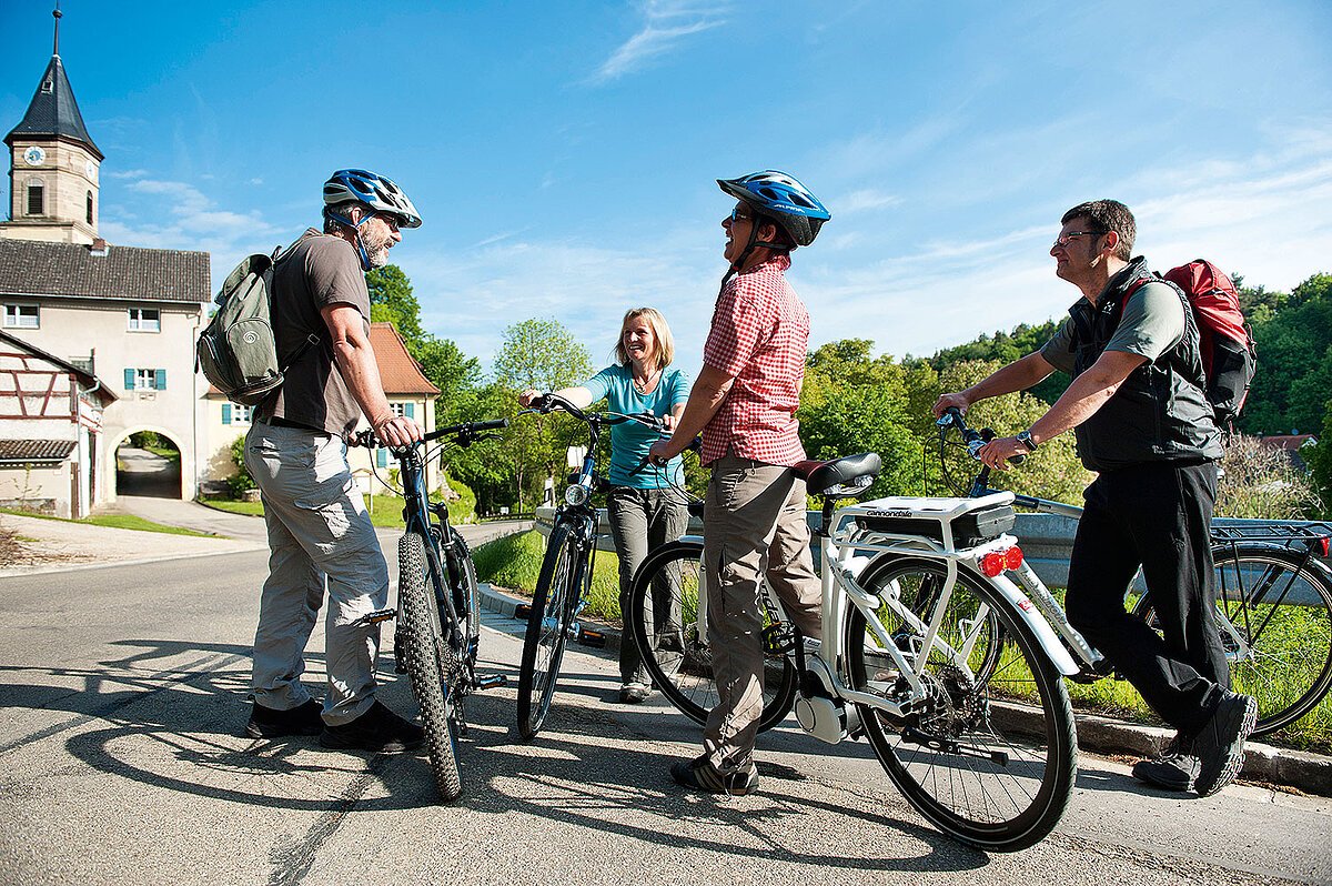 Vier E-Bike Fahrer stehen mit ihren Fahrrädern auf einem geteerten Radweg und unterhalten sich. Im Hintergund stehen Häuser im fränkischen Baustil.