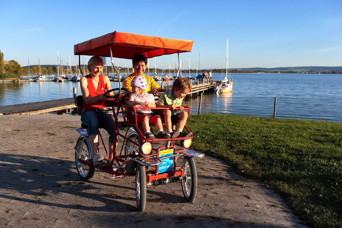 Rikschafahren Eine Ehepaar mit zwei Kindern sitzt in einer Rikscha direkt am Ufer eines Sees. Im Hintergund ein Holzsteg der auf den See führt.